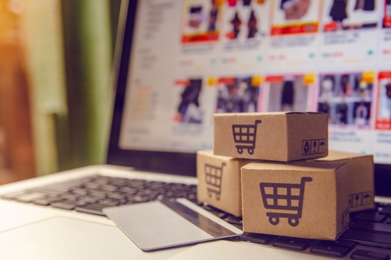 A stack of three cardboards boxes with the image of a shopping cart printed on them, resting on top of a laptop with the screen open to an e-commerce marketplace