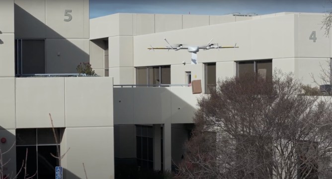 A Wing brand delivery drone flies near a white building with a cardboard box in a promotional video.