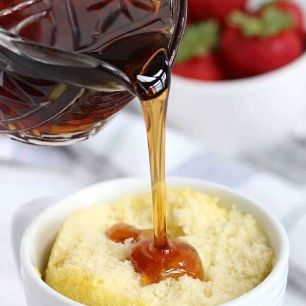 Syrup cascades from a glass jug onto a delightful pancake in a mug, with fresh strawberries adding a splash of color in the background.