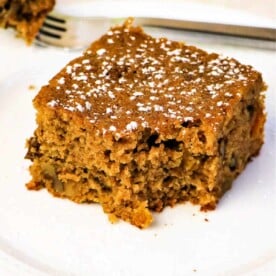 A slice of applesauce spice cake with nuts, dusted with powdered sugar, rests on a white plate, accompanied by a fork in the background.