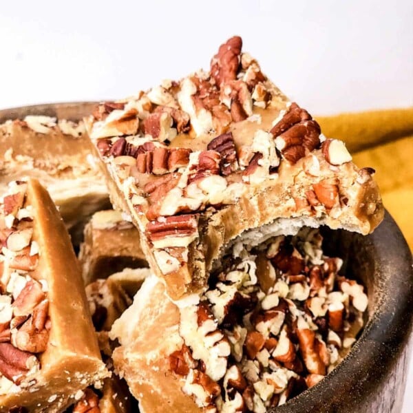 Close-up of a wooden bowl filled with pieces of pecan praline fudge. The pralines, topped with chopped pecans, showcase their rich, caramelized texture. The bowl rests on a white surface with a hint of a yellow cloth in the background.