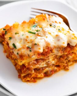 A white plate with Lasagna on it with a fork in the background.