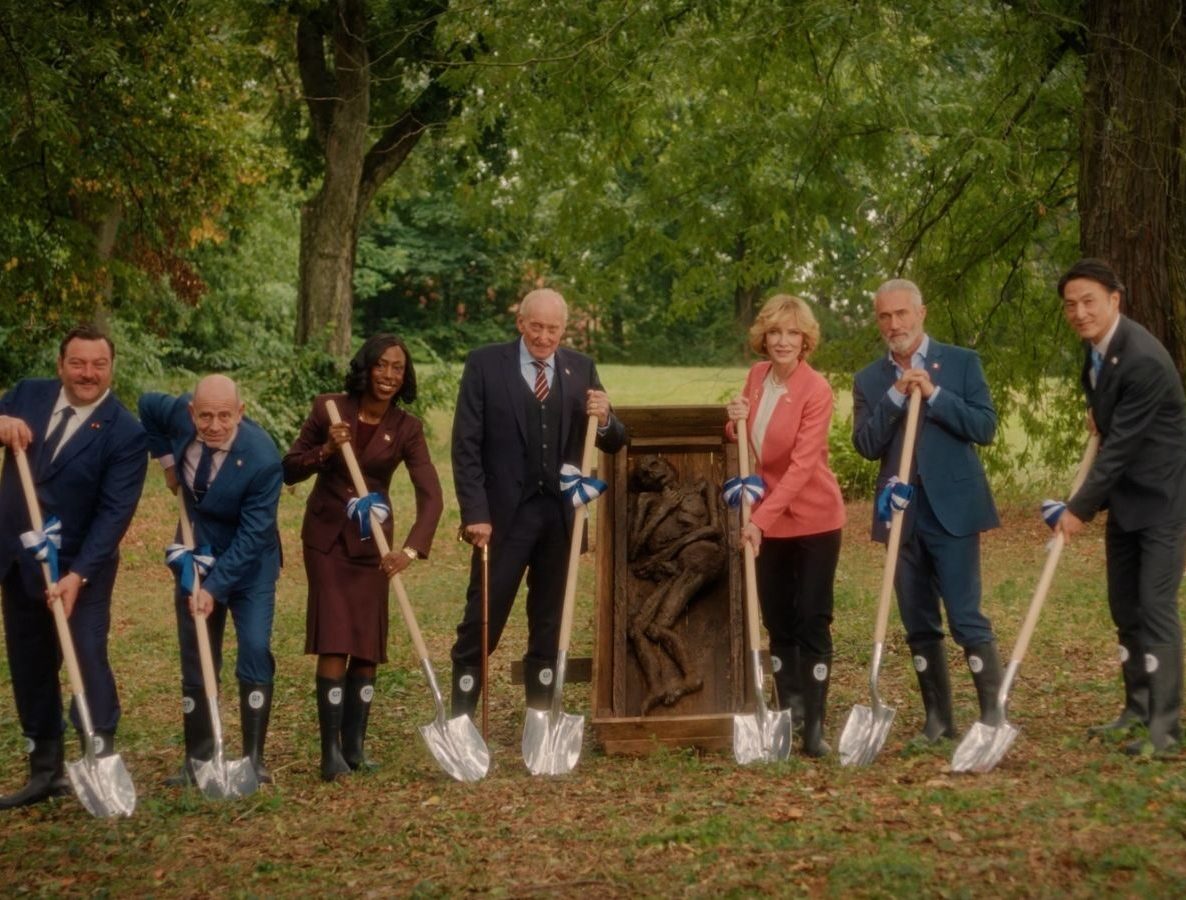 7 people holding shovels over an open grave smiling at camera.