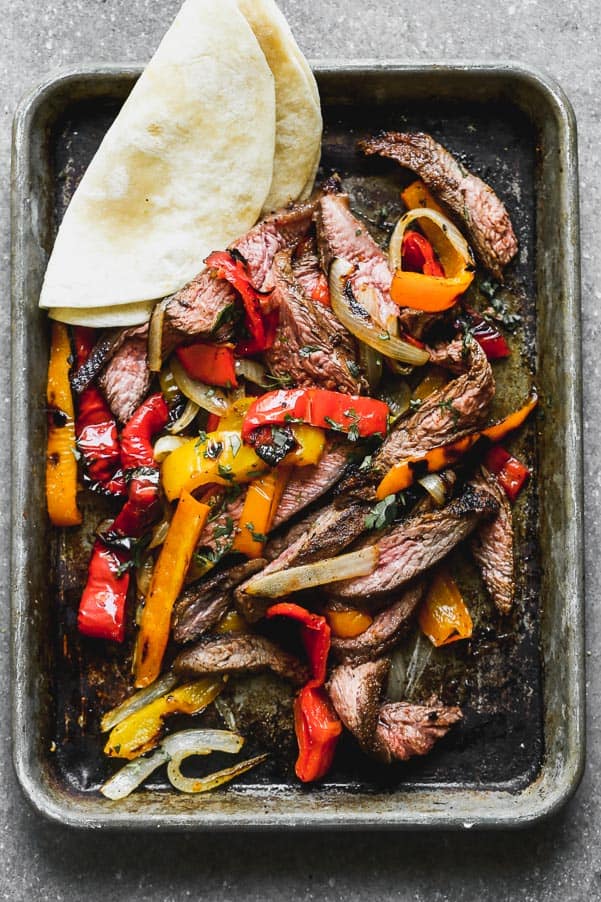 Grilled steak fajitas on a sheet pan with a tortilla on the side. 