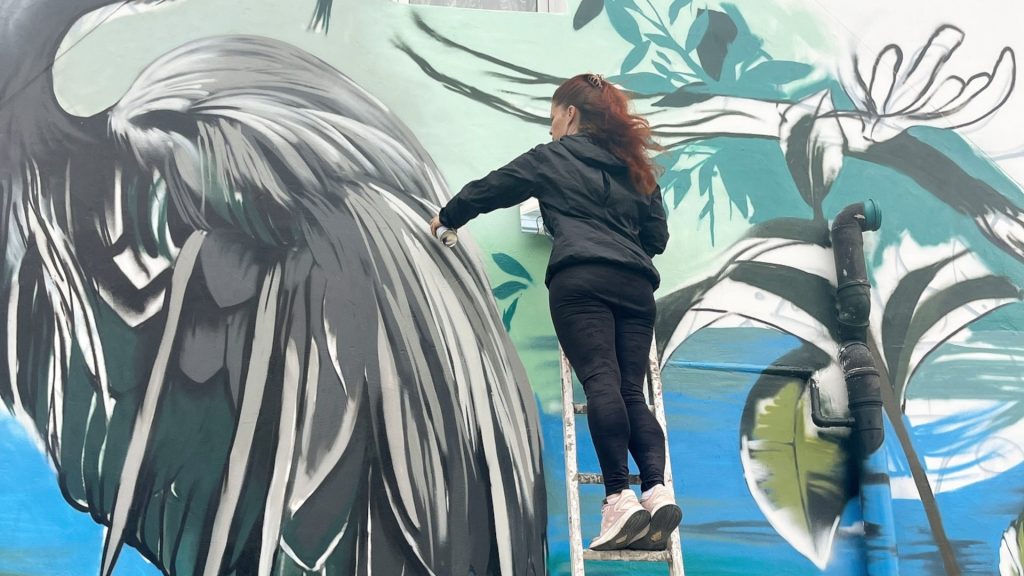 A woman on a ladder adds paint to an enormous image of a bird