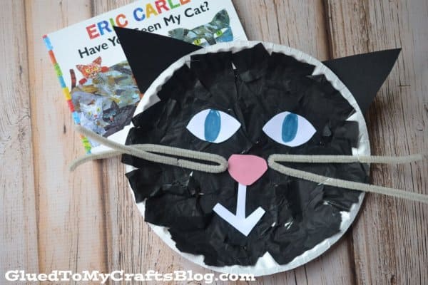 A cat made by gluing black tissue paper to a paper plate with gray pipe cleaner whiskers, with a pink construction nose.