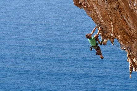 A man climbs up the side of a rock above the ocean.