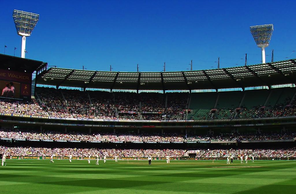 A cricket test match being played at an at capacity Melbourne Cricket Ground on a sunny day. 