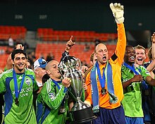 Several players are standing together with one lifting a large trophy upward