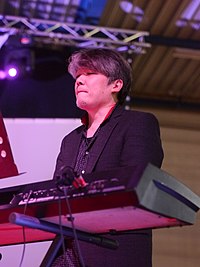 A Japanese man with graying hair stands behind a piano keyboard.