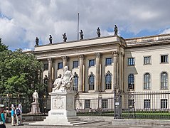 Frontale Farbfotografie von einem klassizistischen Gebäude mit einer großen Statue aus weißem Marmor vor dem Zaun. Das dreigeschossige, helle Gebäude ist mit Säulen und Ornamenten an den Fenstern geschmückt. Auf dem Dach stehen sechs Skulpturen und die Aufschrift „Humboldt Universität“ steht im Balken.