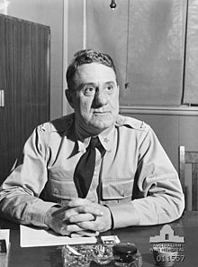 A man wearing a shirt and tie sits at a desk. On the desk are papers and a cigarette in an ash tray.