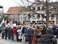 Photographie d'une place publique, avec une foule regardant une estrade