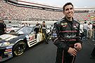 A Hispanic racecar driver dressed in full racing gear poses besides a black race car.