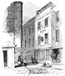 A monochrome illustration of a narrow street, viewed from a corner, or intersection. A large three-storey building is visible on the right of the image. The ground floor has three windows, the first and second floors have two windows each. The roof appears to contain a row of windows, for a loft space. The word "KING" is written between the first and second floors, and a sign, "Gas meter maker" hangs above the ground floor windows.