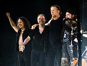 Four men in dark clothing on a stage; the man on the left has his arm raised in the air, while the third man from the left has his arms around the second and fourth.