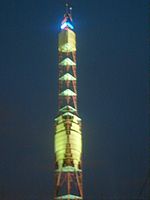 A red lattice tower seen at night with decorative yellow panels in a zig-zag diamond shape and more panels up top. A light panel sits just below the top of the mast and is lit a bright blue.
