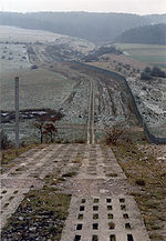 A patrol road, made of two parallel rows of perforated concrete blocks, performs a steep descent into a valley. To the right, running parallel to the road, is a continuous fence. The road and fence continue into the distance, crossing fields that are dusted with a light sprinkling of snow, and ascending another hillside on the far side of valley. Dark forests loom in the distance.