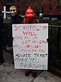 A protester dressed as a Charmeleon from Pokemon holds a sign parodying Rick Astley's "Never Gonna Give You Up" and the "Rickroll" meme while making references to Scientology's tax-exempt status and their "Fair Game" policy