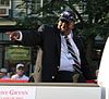 Profile view of a dark-skinned, heavyset man wearing a dark suit, white dress shirt, and a ballcap. He is in an elevated, sitting position in a car pointing to the left with his right arm.