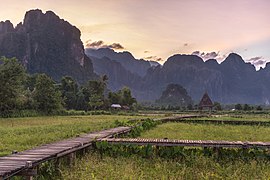 Wooden walkway leading to a hut with straw roof in front of karst mountains at sunset, Vang Vieng, Laos