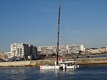 Voilier blanc s'approchant au moteur d'un quai. En arrière-plan, grands immeubles.