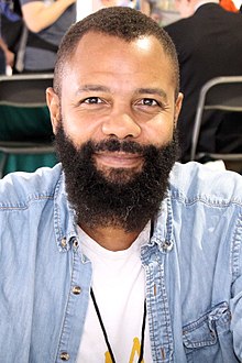 A headshot of Javaka Steptoe smiling at the camera. He has short hair and a beard, and wears a blue jean shirt over a white t-shirt.