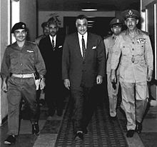 Three important men walking in a hall, the first and the third are in military garb, the second is in a suit and tie. Behind them are three other men