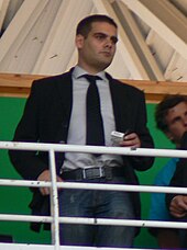 Le portrait d'un homme en costume cravate présent dans les tribunes d'un stade de rugby.