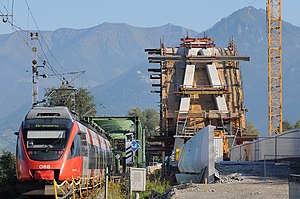 Red train crosses old green bridge as new arched bridge is constructed at right