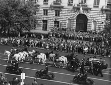 A black-and-white photograph of a wagon being drawn and accompanied by seven white horses, some of which are being ridden by people