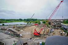 On an overcast day, two cranes and several other construction vehicles work in a mostly empty worksite between some one-story buildings, a road, and a lake.