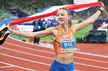 Photo of Femke Bol holding the Dutch flag over her head with the tracks in the background