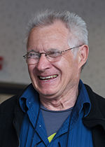 A older man is seen wearing a black coat over a royal blue collared shirt.