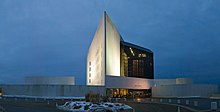 A white triangular tower rises beside a black glass building, with circular structures on either side.