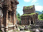 Ruins of buildings of red stone with niches and sculptures. The roof of one of the structures is partially covered in grass.