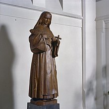 Wooden carving of a full length nun standing holding a large quill pen in her right hand and a crucifix in her left