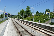 In der Bildmitte verlaufen zwei parallele Gleise, links und rechts davon jeweils ein typischer S-Bahnsteig mit Schildern, Uhr, Mülleimer etc. Hinter den Bahnsteigen sieht man jeweils eine begrünte Umgebung.