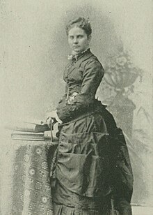 B&W photo of a standing woman with her hair in an updo, wearing a dark dress.