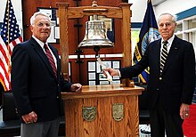 An interior wooden display case with a brass ship's bell flanked by two men in coat and tie standing on either side.