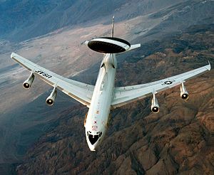 Aerial view port view of white jet aircraft in-flight. It has a large disc-like black radar lying horizontally above two convergent struts.