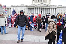 A QAnon emblem (upper left) is raised during the 2021 United States Capitol attack. 102.East.USCapitol.WDC.6January2021 (50824307713).jpg