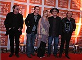 Five people (four men, one woman in the middle) standing next to one another on a red carpet; in the background is a repeated pattern of orange blocks with the text "meteor IRELAND MUSIC AWARDS"