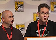 A man with a bald head and a brown sweater, and a man with spiked brown hair and glasses, speaking into a microphone.