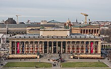 Frontale Farbfotografie eines dunklen Gebäudes mit einer durchgehenden Säulenvorhalle, hinter der sich ein breiter Glaseingang und rote Kachelwände befinden. Ein lateinischer Text steht im Gesims. Ein rechteckiger Aufbau mit Pferden- und Menschenfiguren sowie einer Fahne befindet sich auf der Dachmitte. Im Vordergrund ist eine Wiese mit einem Springbrunnen.