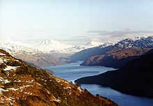 In the distance there are high snow-capped mountains, out of which a sinuous lake emerges. In the foreground lower brown hills surround the water.