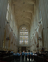 Interior of large building with a stained glass window at the far end. Above is a barrel vault ceiling and on either side rows of arches. People standing at the bottom of the picture dwarfed by the height of the ceiling.