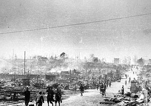 Black and white photo of people walking along a road passing through a large area of destroyed buildings