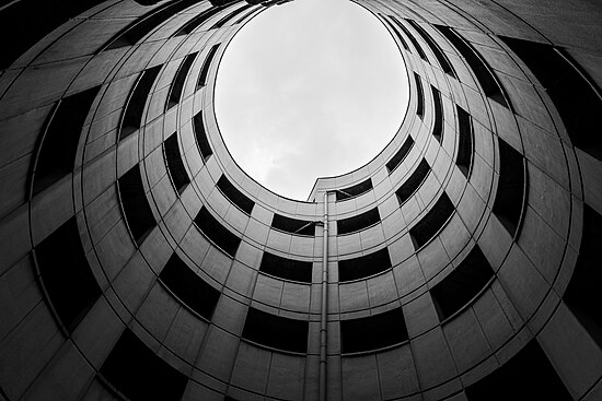 Senkrechter Blick nach oben in eine Parkhaus-Auffahrt in S/W, darüber ein grauer Himmel