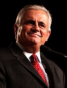 A Caucasian male wearing a black suit with a white dress shirt and red patterned tie is seen against a black background.